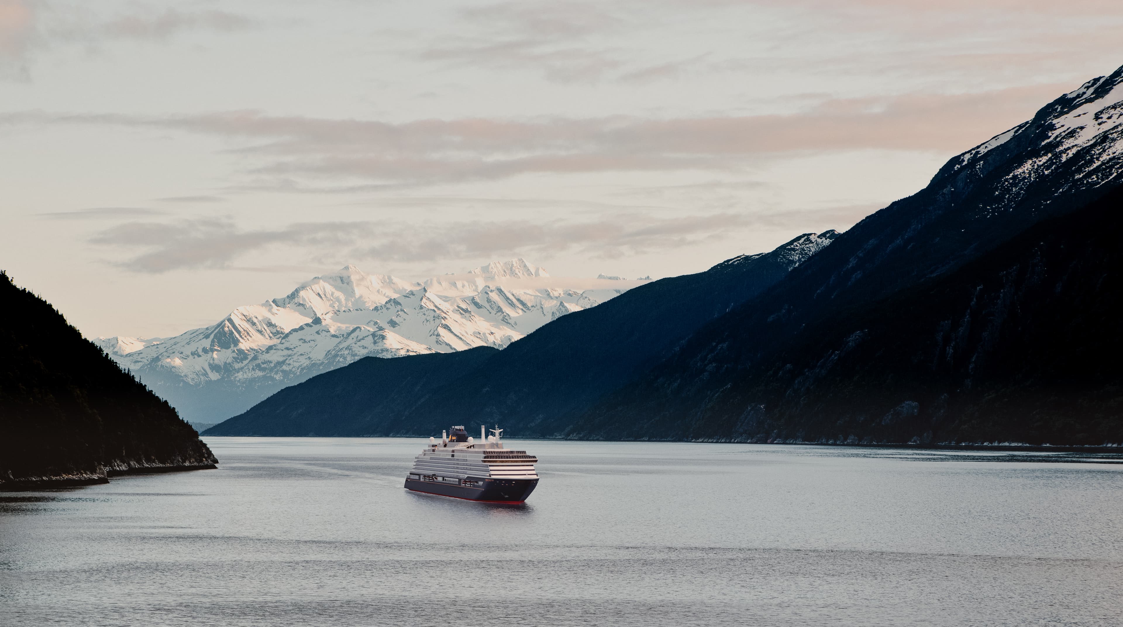 Explora Journeys ship sailing through an Alaskan fjord at dusk, surrounded by snow-capped mountains reflected in calm water