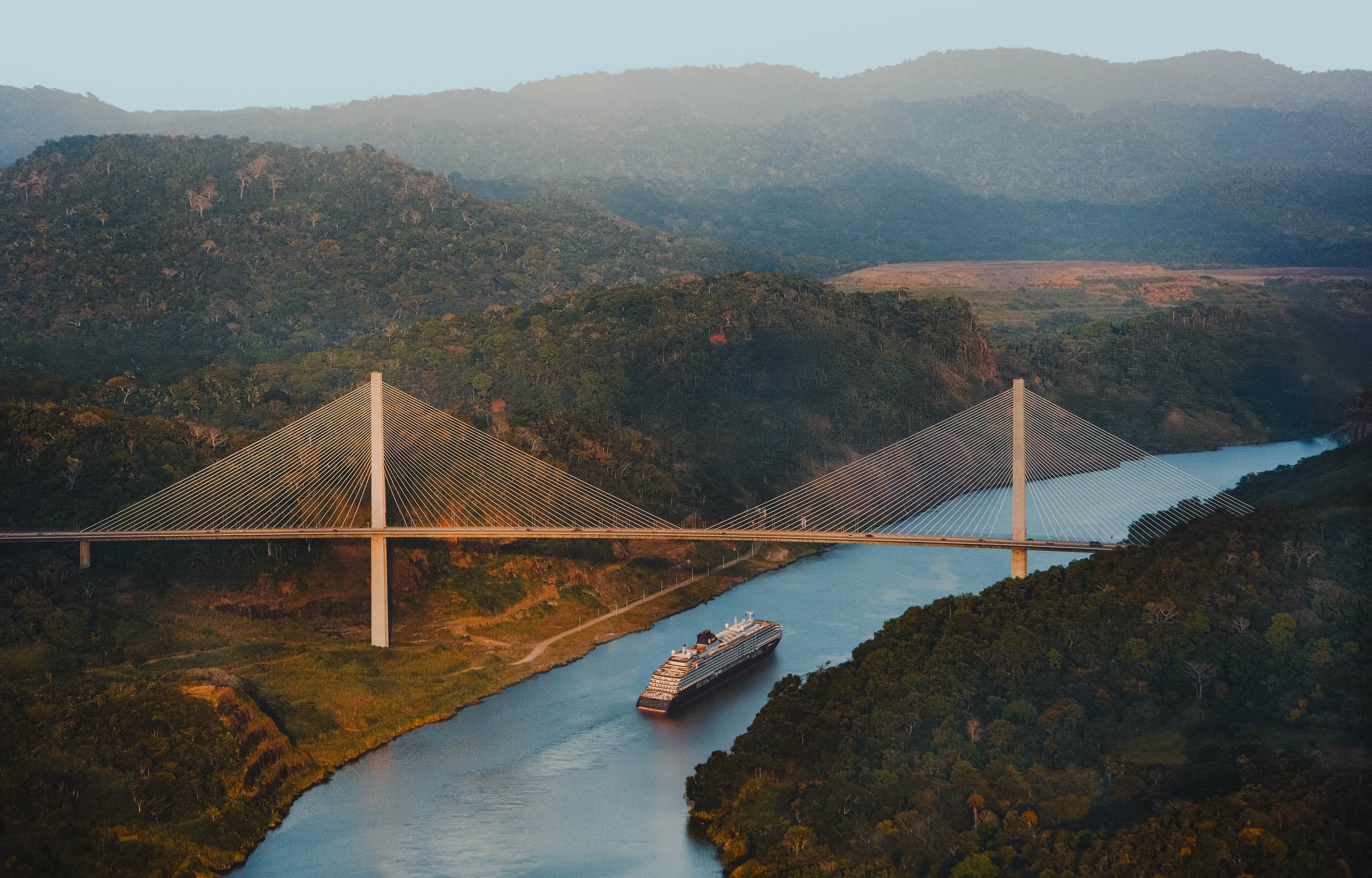 Explora Journeys ship transiting the Panama Canal beneath the Centennial Bridge, surrounded by lush tropical jungle at golden hour